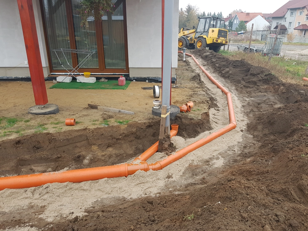 Trwa montaż drenażu opaskowego – pomarańczowe rury osadzone w obsypce piaskowej wokół fundamentu domu