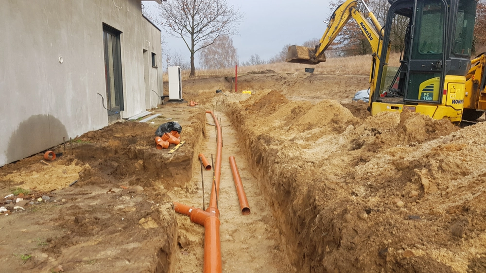 Wykop pod drenaż opaskowy z ułożonymi pomarańczowymi rurami i mini koparką przy budynku