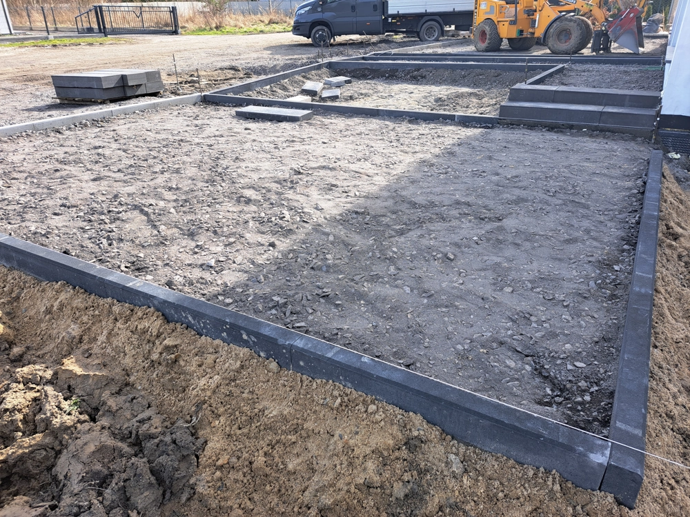 Układanie krawężników betonowych na wyrównanym podłożu placu budowy z widocznym zagęszczonym gruntowiskiem