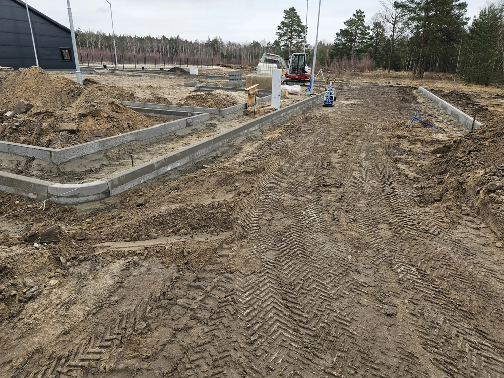 Niwelacja terenu na placu budowy z widocznymi śladami ciężkiego sprzętu i osadzonymi krawężnikami betonowymi