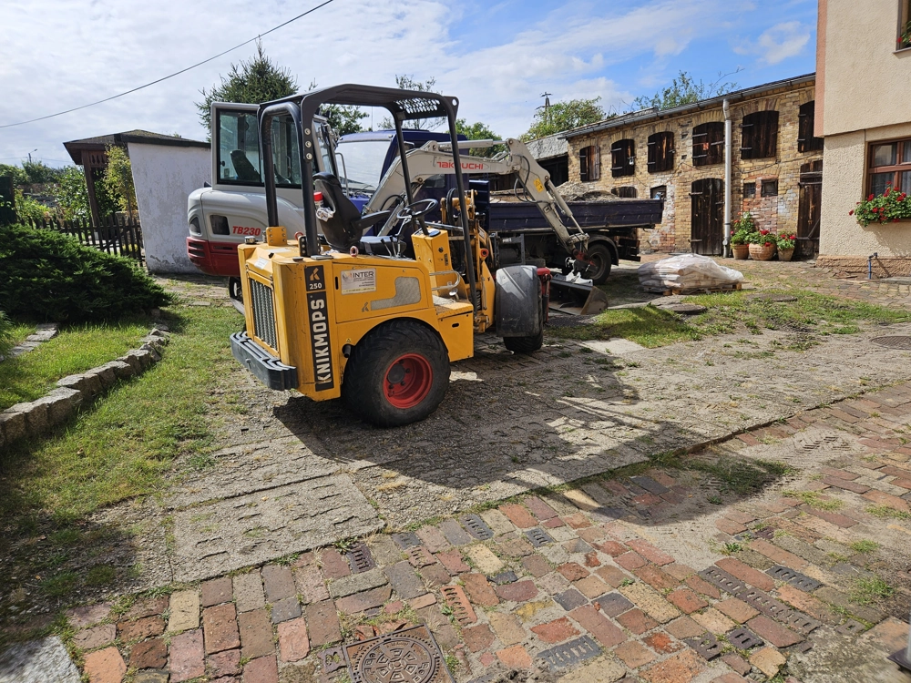 Wozidło budowlane na brukowanym podwórzu obok minikoparki