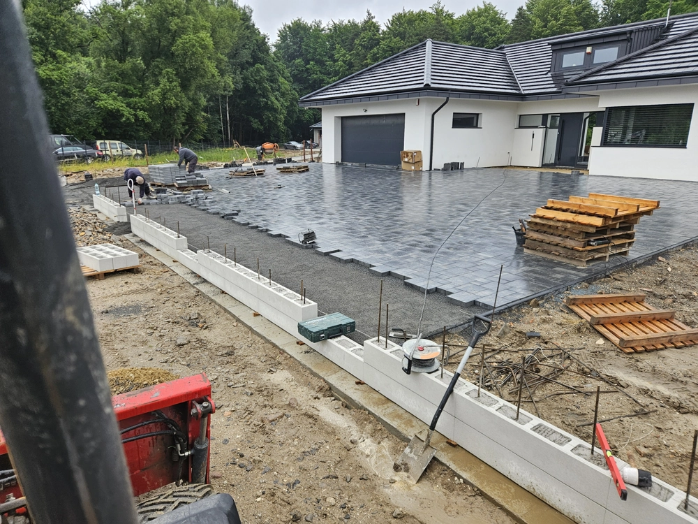 Brukarze układają ciemnoszare kostki brukowe na podbudowie podjazdu przy nowoczesnym domu