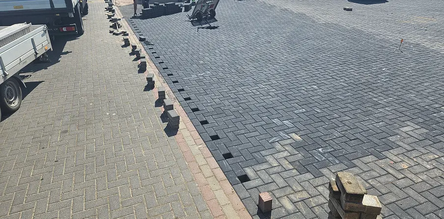 Układanie kostki brukowej w układzie jodełki na podjeździe, widoczne przerwy przy krawędziach i stosy kostki
